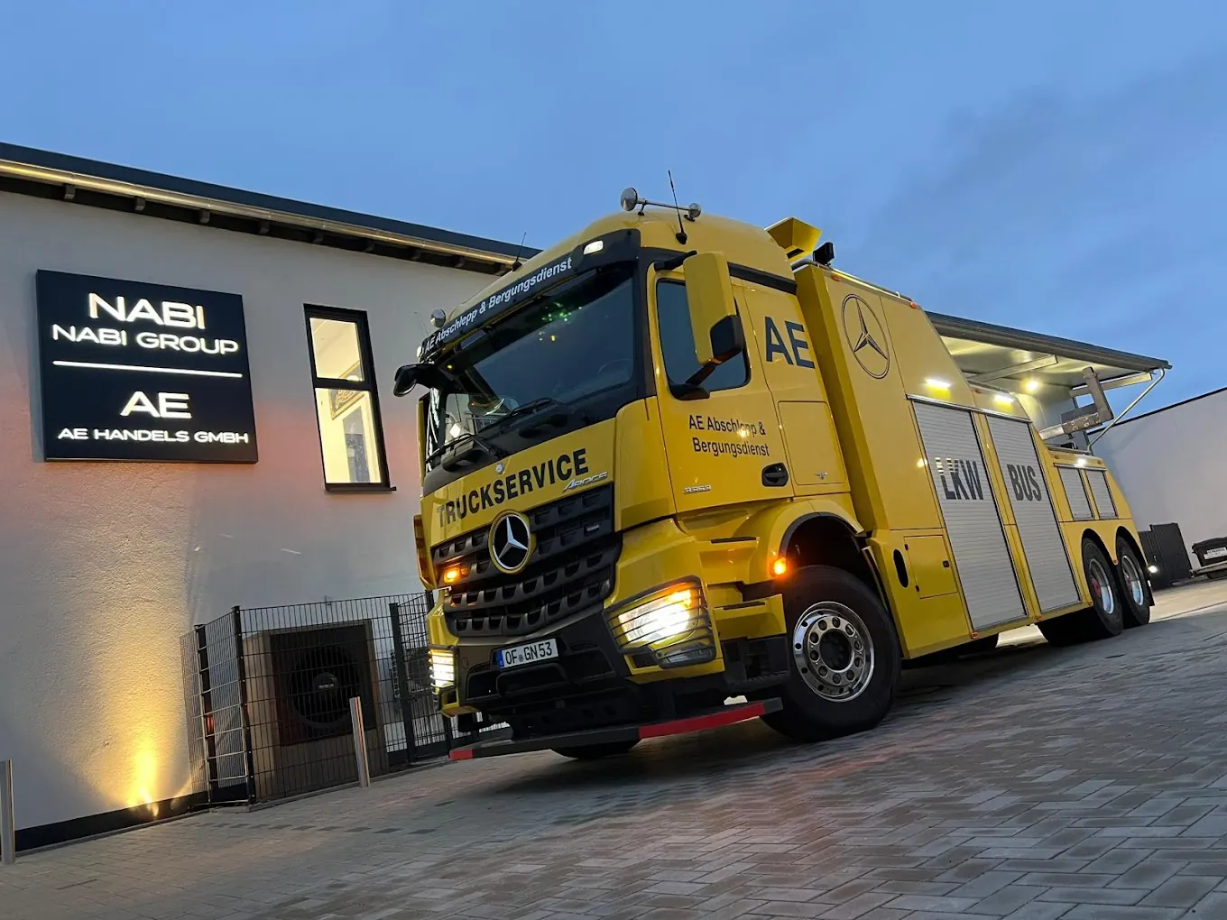 Gelber Abschlepp-Lkw von AE vor dem beleuchteten Firmengebäude in Mühlheim am Main